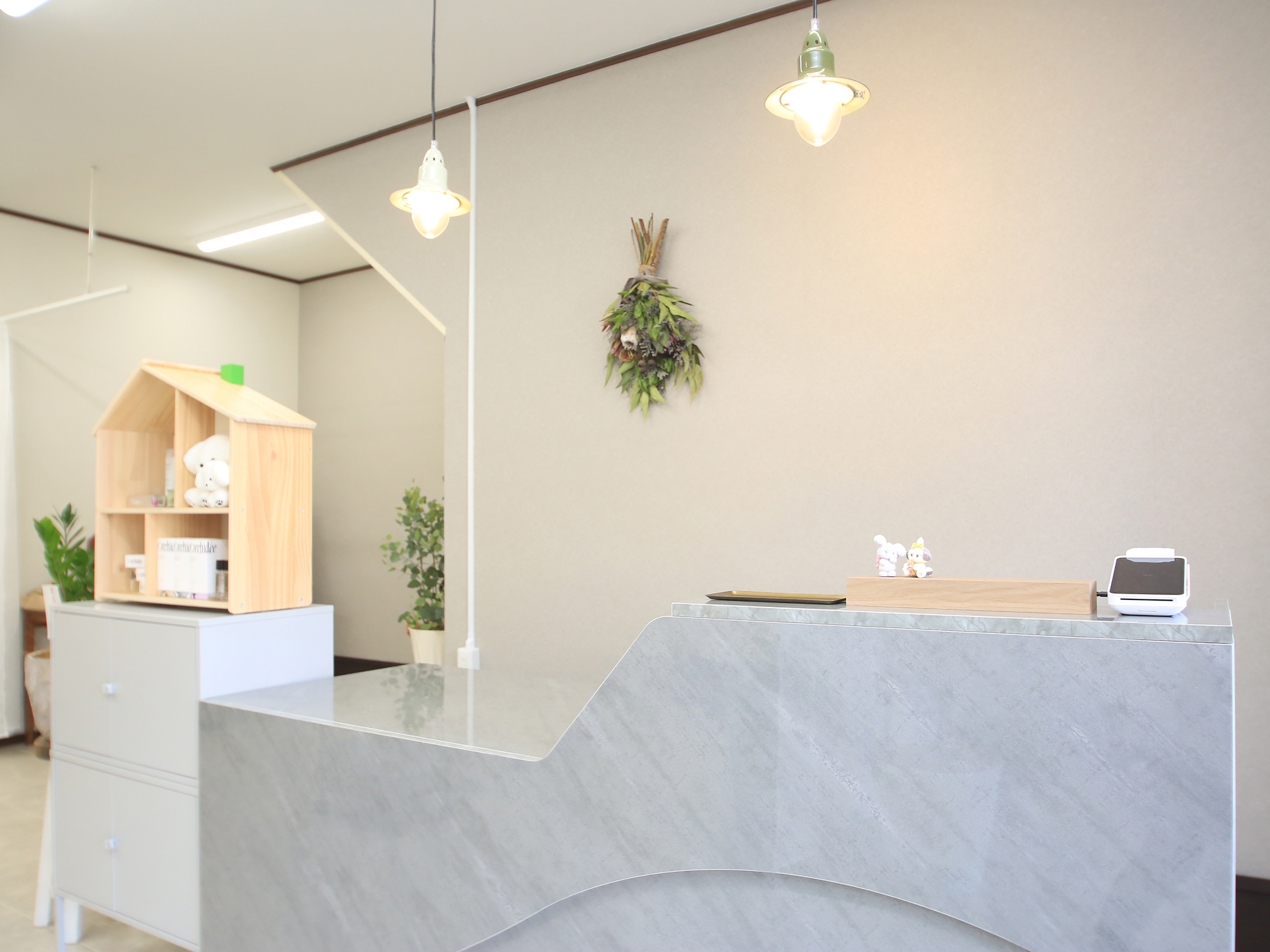 アイラッシュサロン経営者の施術室 - Growth Partners Japan導入事例 アイラッシュサロン受付内観 白とグレーの曲線的な受付カウンター ナチュラルな照明と植物装飾 清潔で温かみのある空間 関東 30代女性オーナー経営